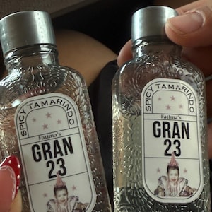 May include: Two clear glass bottles with silver caps. Each bottle has a label that reads "Spicy Tamarindo Fatima's GRAN 23" with a picture of a baby. The bottles are held by a person.