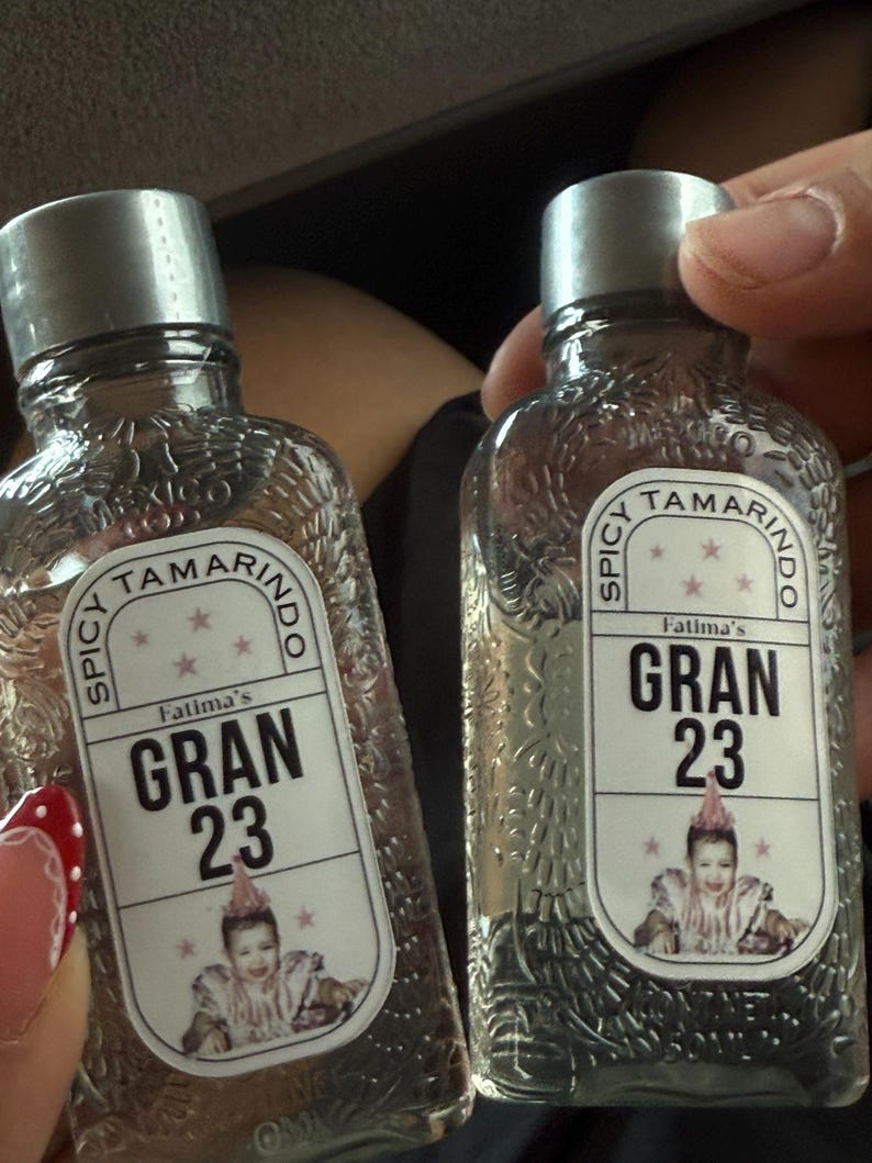 May include: Two clear glass bottles with silver caps. Each bottle has a label that reads "Spicy Tamarindo Fatima's GRAN 23" with a picture of a baby. The bottles are held by a person.