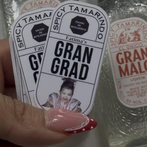 May include: A stack of stickers with the text "Fatima's Gran Grad" and a photo of a person in a graduation cap. The stickers are held in front of two bottles of "Spicy Tamarindo" liqueur.
