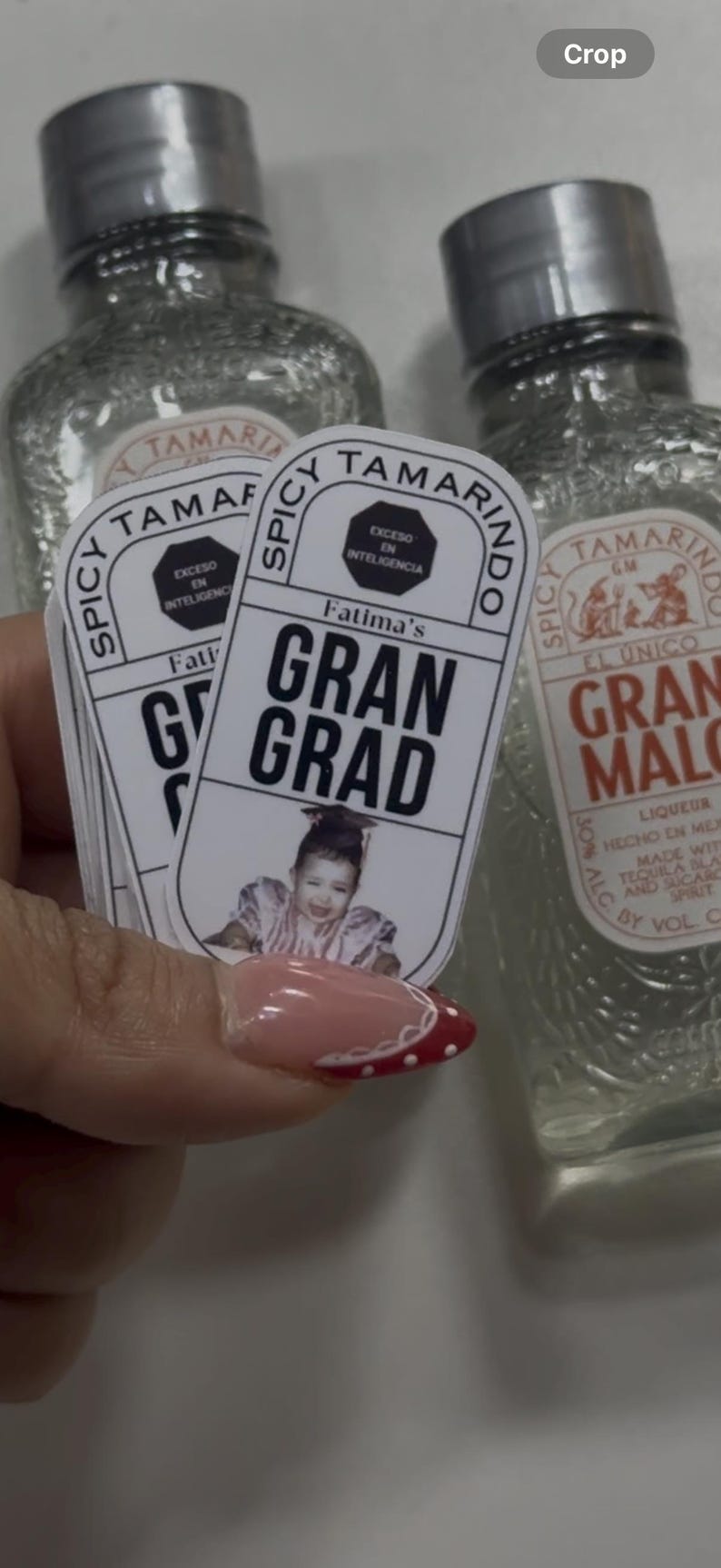 May include: A stack of stickers with the text "Fatima's Gran Grad" and a photo of a person in a graduation cap. The stickers are held in front of two bottles of "Spicy Tamarindo" liqueur.