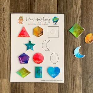 May include: A colorful educational shape matching puzzle for kids. The puzzle features a variety of shapes, including a triangle, square, circle, star, heart, hexagon, oval, crescent moon, diamond, rectangle, and pentagon. Each shape is labeled with its name.