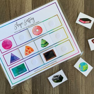 May include: A shape sorting educational game with a white background and a rainbow border. The game includes shapes like circles, triangles, and rectangles, with matching picture cards of a donut, pizza, and Christmas tree. The text "Shape Sorting" is visible.