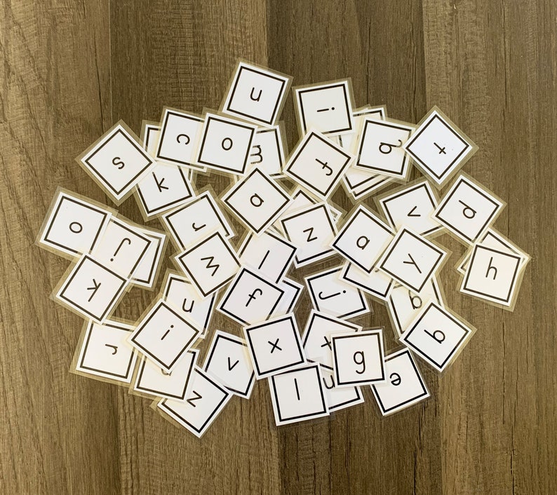 K&ouml;nnte beinhalten: Eine Sammlung wei&szlig;er Buchstabenkacheln mit schwarzer Beschriftung, verstreut auf einer Holzoberfl&auml;che. Die Buchstaben umfassen: a, b, c, d, e, f, g, h, i, j, k, l, m, n, o, p, q, r, s, t, u, v, w, x, y, z.