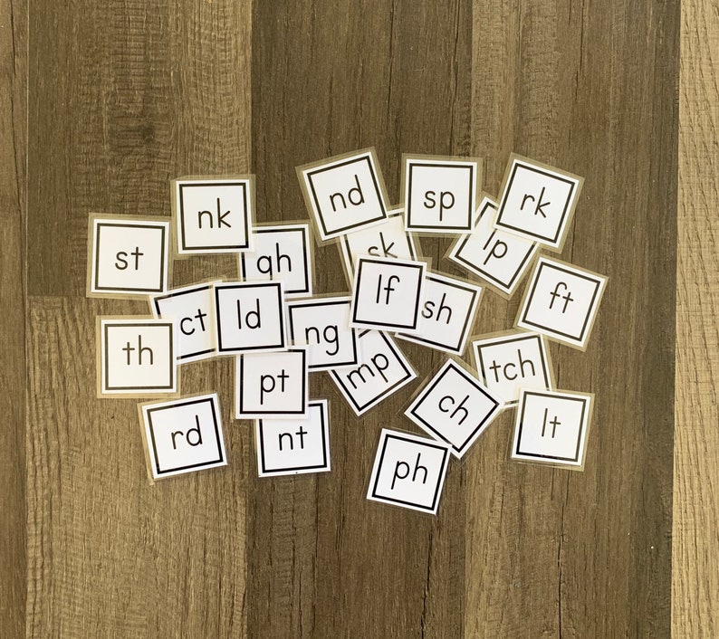K&ouml;nnte beinhalten: Ein Satz von 20 wei&szlig;en quadratischen Karten mit schwarzem Text, jede Karte zeigt eine andere Zwei-Buchstaben-Kombination. Die Karten sind auf einer Holzoberfl&auml;che verstreut.
