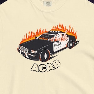 May include: Cream-colored t-shirt featuring a graphic of a burning police car with the word "POLICE" on the side and "ACAB" below. The car is black with orange flames and a cartoon character driving.