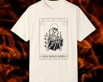 May include: White t-shirt with a black and white illustration of a skeleton reading a book. The illustration is in the style of a tarot card and includes the text "XVIII" and "Read Banned Books".