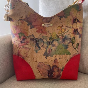 May include: A cork handbag with a floral print in shades of red, yellow, and green. The bag has a red leather trim and a gold metal tag that reads "GRAND handbags".