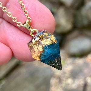 Op de afbeelding: Een ketting met hanger bestaande uit een gefacetteerde, geometrische harsbedel aan een goudkleurige ketting. De bedel bevat een mix van diepblauwe hars, ingebedde bladgoudvlokken en een donkere, getextureerde basis met kleine metaaldeeltjes.
