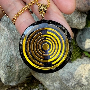 May include: A round black pendant featuring a gold-coloured concentric circle pattern on its surface. The pendant is suspended from a gold-coloured metal chain. The dark material of the pendant contains small, reflective metallic flecks throughout.