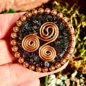 May include: A circular pendant featuring a dark, textured, glittery black base embedded with a central copper wire triskelion design. The outer edge of the disc is decorated with a continuous border of small, polished copper-coloured metallic beads.
