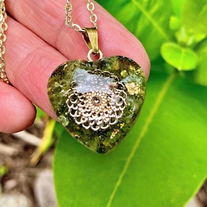 May include: A heart-shaped pendant necklace featuring a translucent olive green resin base embedded with gold-colored metallic flakes and a central circular filigree charm. The pendant hangs from a gold-toned metal chain link necklace.