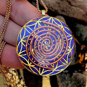 May include: A round pendant necklace featuring a deep blue background with a gold-colored geometric flower of life pattern. A copper-colored metal wire is coiled in a spiral shape on the front surface. The pendant hangs from a gold-colored chain.