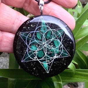 Peut inclure: Pendentif rond avec un fond noir parsemé de paillettes dorées. Le pendentif présente un motif géométrique blanc et des pierres de malachite vertes. Il est suspendu à un cordon noir avec un fermoir argenté.