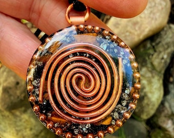May include: A circular pendant featuring a central copper wire spiral encased in clear resin. The interior contains embedded dark metallic flakes and amber-toned fragments. The pendant is framed by a beaded copper border and a top bail.