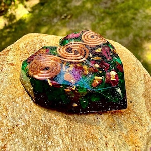 May include: A heart-shaped resin object containing embedded green stone fragments, deep red petals, and gold leaf flakes. Three circular copper wire coils are arranged in a line across the top surface of the translucent, multi-coloured piece.