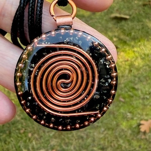 May include: A circular pendant featuring a central copper wire spiral embedded in a black resin base with metallic flecks. The pendant is framed by a border of small copper beads and hangs from a black cord by a copper bail at the top.