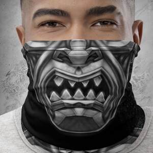 May include: A black and grey face covering featuring a detailed illustration of a samurai mask. The mask design includes sharp teeth and intricate armour details. The face covering is worn by a person wearing a grey shirt. The logo "MONSTRO INT." is visible.