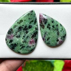 Może przedstawiać: Dwa wypolerowane kamienie szlachetne w kształcie łezki. Kamienie mają zielony kolor bazowy z czarnymi i różowymi wtrąceniami. Kamienie szlachetne są prezentowane na białej powierzchni z rozmytym czerwonym i zielonym tłem.