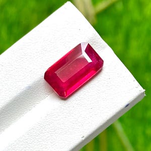 May include: A vibrant, deep red gemstone cut into a rectangular emerald shape with faceted surfaces. The stone displays a translucent quality and sits on a white surface, showcasing its sharp edges and polished, reflective appearance.