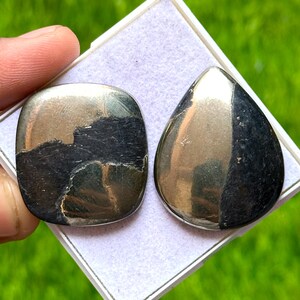 Pode incluir: Duas pedras preciosas de pirita polidas. Uma é quadrada com cantos arredondados, a outra em forma de gota. Ambas têm uma cor dourada e preta metálica, com uma superfície refletora. As pedras são exibidas numa bandeja branca sobre um fundo verde.