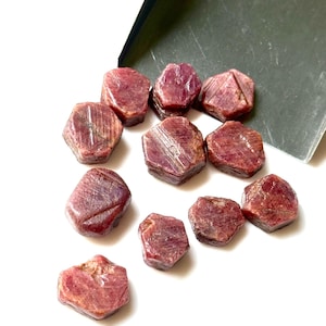 Op de afbeelding: Een verzameling ruw geslepen, dieprode robijn edelstenen verspreid op een wit oppervlak. De edelstenen hebben een natuurlijke, ongepolijste uitstraling, met zichtbare texturen en facetten. Een donkergroene kaart met de tekst "CHB NO. 3" staat op de achtergrond.