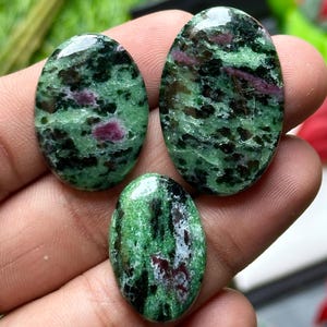 May include: Three oval-shaped gemstones with a mix of green, black, and red hues. The stones have a polished, glossy surface, and the colors are swirled and speckled throughout. The gemstones are likely used for jewelry or decorative purposes.