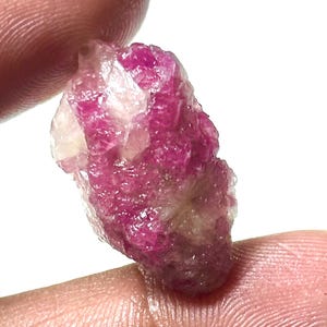 Könnte beinhalten: Ein roher, ungeschliffener Edelstein in leuchtendem Rosa, gehalten zwischen Fingern vor weißem Hintergrund. Der Stein weist eine kristalline Struktur mit helleren, fast weißen Bereichen auf. Die Gesamtform ist unregelmäßig und natürlich.
