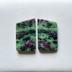 May include: Two polished ruby zoisite stones. The stones are rectangular with rounded corners and a slight trapezoid shape. The stones have a green base color with black and pink inclusions. The stones are displayed on a white surface.