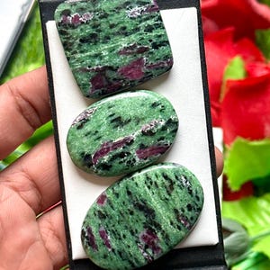 May include: Three polished zoisite stones with ruby inclusions. The stones are green with black and red marbling, and are rectangular and oval shaped. The stones are displayed on a white surface.