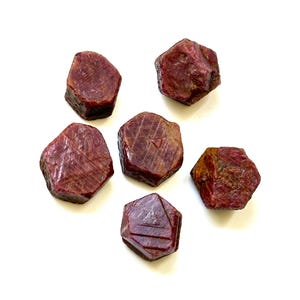 Puede incluir: Seis piedras preciosas de rubí rojo oscuro en bruto se muestran sobre un fondo blanco. Las piedras tienen un aspecto natural, sin pulir, con texturas y facetas visibles. Los rubíes varían ligeramente en forma y tamaño.