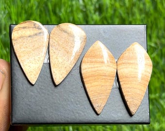 May include: Four polished, teardrop-shaped gemstones with a warm, beige color and natural veining. The stones are displayed on a black surface against a backdrop of green grass. These stones are ideal for jewelry making.