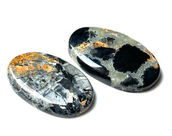 Puede incluir: Dos piedras preciosas ovaladas pulidas. Las piedras presentan una mezcla de colores negro, gris y dorado. Las piedras preciosas tienen un acabado brillante y están sobre un fondo blanco. Estas piedras son ideales para la fabricación de joyas.