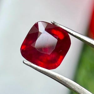 May include: A vibrant, cushion-cut ruby gemstone held by tweezers. The ruby displays a deep red hue with multiple facets, reflecting light. The gemstone is the focal point, showcasing its clarity and rich color. The tweezers are silver.