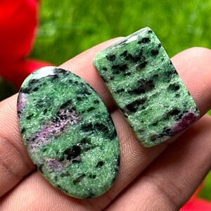 May include: Two polished zoisite gemstones. One is oval-shaped, the other rectangular. Both feature a green base color with black and pink inclusions. The gemstones are held in a hand against a blurred green background.