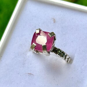 May include: A silver ring with a large, square-cut, pink gemstone. The ring's band features a detailed floral pattern. The gemstone is set in a silver bezel, and the ring is displayed in a white box against a blurred green background.
