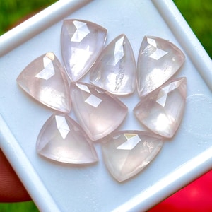 May include: A collection of seven pale pink, triangular-shaped gemstones with faceted surfaces arranged in a white display tray. Each stone features a translucent appearance and a polished finish, reflecting light across the multiple cut geometric planes.