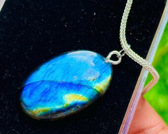 May include: Oval labradorite pendant with a silver chain. The gemstone displays a vibrant blue color with yellow and green flashes. The pendant is set in a silver bail and is displayed on a black velvet surface.