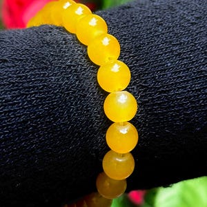 May include: A beaded bracelet featuring round, polished yellow beads. The beads are strung together, creating a flexible band. The bracelet is displayed against a dark background, highlighting the vibrant color of the beads.