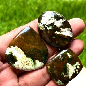 May include: Three polished gemstones in various shapes: a teardrop, an oval, and a square. The stones feature a mix of brown, green, and white colors, with the white areas appearing textured. The gemstones are held in a hand against a blurred green background.