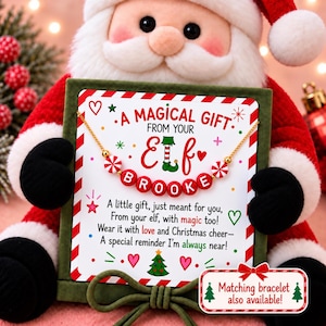 May include: A gold-tone chain necklace featuring red and white peppermint-style beads and red letter beads spelling BROOKE, displayed on a card with Christmas text and graphics, held by a plush Santa Claus figure.