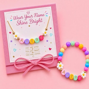 May include: A gold-tone chain necklace and a beaded bracelet, both featuring colorful letter beads spelling AVERY and white daisy charms. They are presented on a pink card with the text Wear Your Name Shine Bright and You Are Kind, You Are Smart, You Are Loved, decorated with hearts and stars.