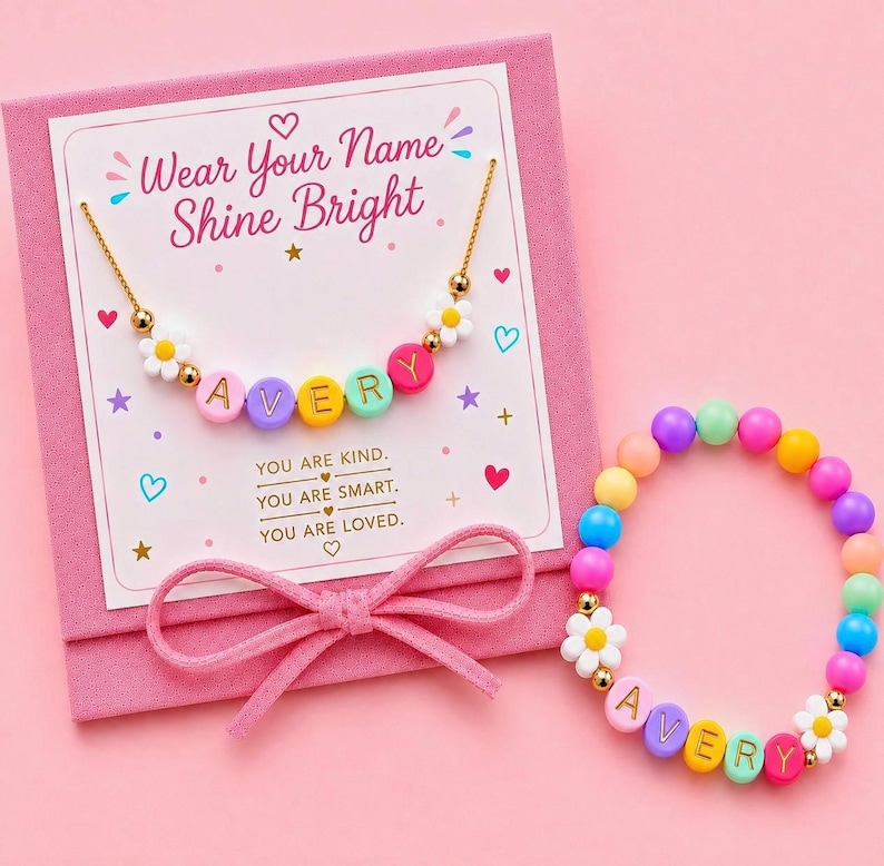 May include: A gold-tone chain necklace and a beaded bracelet, both featuring colorful letter beads spelling AVERY and white daisy charms. They are presented on a pink card with the text Wear Your Name Shine Bright and You Are Kind, You Are Smart, You Are Loved, decorated with hearts and stars.