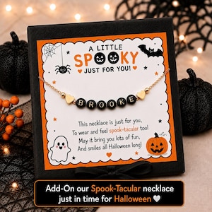 May include: A gold-tone chain necklace featuring black letter beads spelling BROOKE, flanked by two small gold heart charms. The necklace is displayed on a white card with orange borders, decorated with Halloween icons and the text A LITTLE SPOOKY.