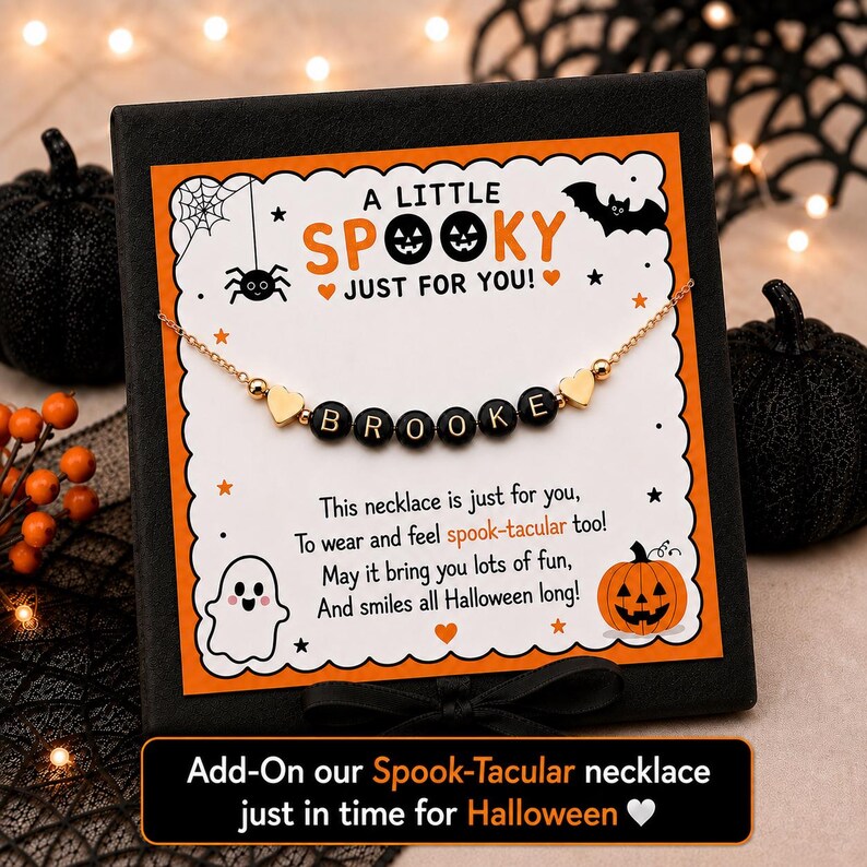 May include: A gold-tone chain necklace featuring black letter beads spelling BROOKE, flanked by two small gold heart charms. The necklace is displayed on a white card with orange borders, decorated with Halloween icons and the text A LITTLE SPOOKY.