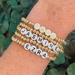 May include: A set of four gold bead bracelets with white beads that spell out the names "Mama", "Jasmine", and "Ezra".