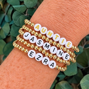 Pu&ograve; includere: Una pila di quattro braccialetti elastici realizzati con piccole perline metalliche dorate. Ogni braccialetto presenta perline bianche rotonde con lettere che compongono AUNTIE in giallo, e JASMINE ed EZRA in nero, tra le perline dorate.