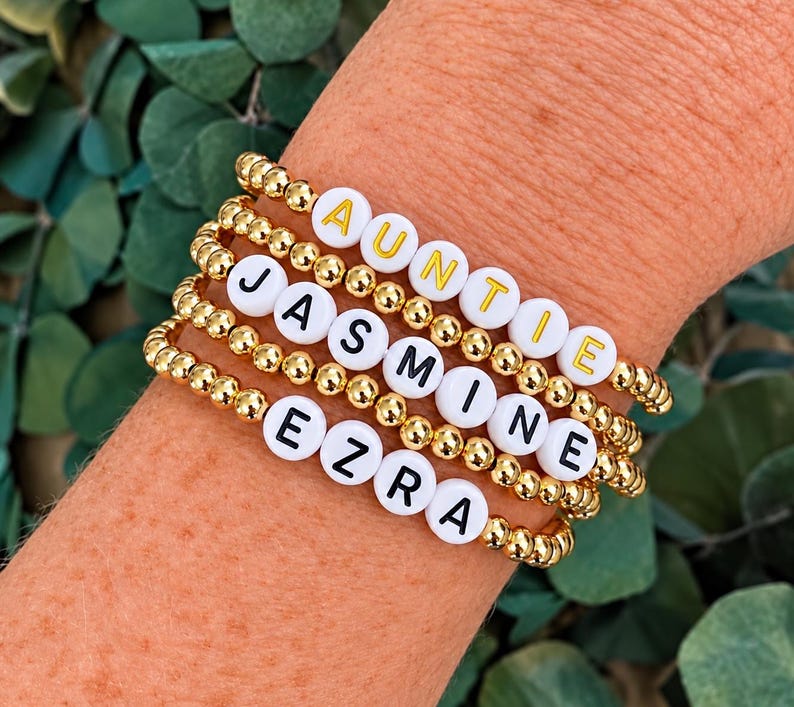Pu&ograve; includere: Una pila di quattro braccialetti elastici realizzati con piccole perline metalliche dorate. Ogni braccialetto presenta perline bianche rotonde con lettere che compongono AUNTIE in giallo, e JASMINE ed EZRA in nero, tra le perline dorate.