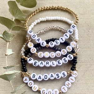 Pu&ograve; includere: Una pila di cinque braccialetti di perline con perle dorate, bianche, trasparenti e nere. I braccialetti includono perline con lettere bianche che compongono le frasi GOAL GETTER, NO EXCUSES, FEARLESS, BADASS e YOU GOT THIS in nero e oro.