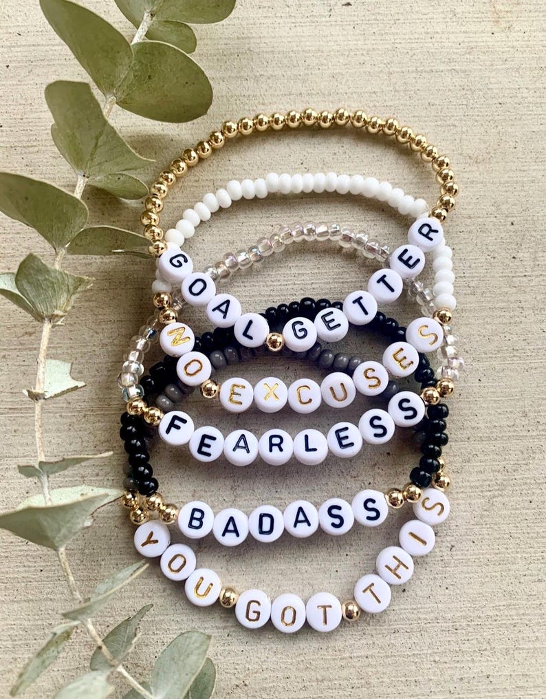 Pu&ograve; includere: Una pila di cinque braccialetti di perline con perle dorate, bianche, trasparenti e nere. I braccialetti includono perline con lettere bianche che compongono le frasi GOAL GETTER, NO EXCUSES, FEARLESS, BADASS e YOU GOT THIS in nero e oro.