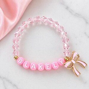 May include: A beaded bracelet featuring translucent light pink faceted crystal beads and round pink letter beads spelling the name BLAKELY. The bracelet includes a gold-toned metal charm shaped like a bow with light pink enamel detailing.